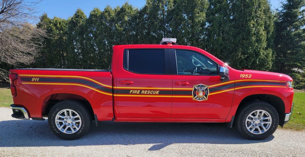 A red pickup truck marked "FIRE RESCUE" from the Liberty Township Volunteer Fire Department, with yellow and black stripes, emergency lights, and the numbers 911 and 1952, is parked on a gravel surface with trees in the background.