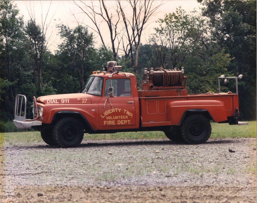 A red vintage pickup fire truck labeled "Liberty Township Volunteer Fire Department" is parked on gravel, with green trees in the background. A large hose reel sits atop the truck bed.