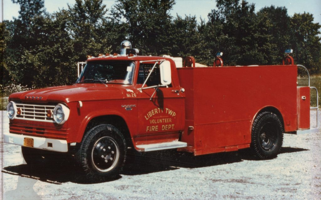 A vintage red fire truck labeled "Liberty Township Volunteer Fire Department" is parked on gravel, surrounded by trees and lush greenery in the background.