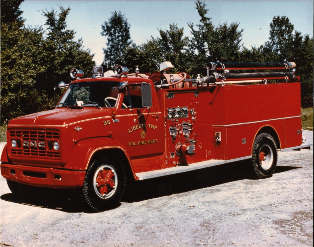 A vintage red GMC fire truck labeled "Liberty Township Volunteer Fire Department" is parked on a gravel surface, with trees and greenery in the background. Firefighting equipment lines the truck’s sides and roof.