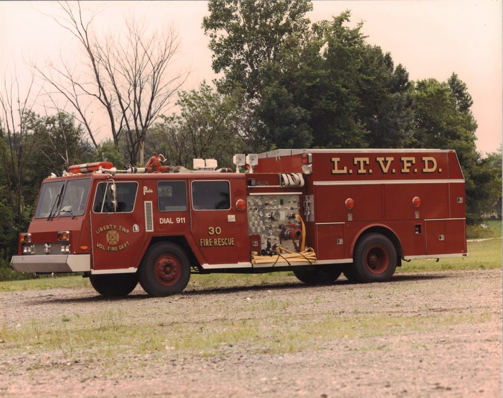 A red fire truck with "L.T.V.F.D." and "Liberty Twp. Vol. Fire Dept." on the side, representing the Liberty Township Volunteer Fire Department, is parked on gravel amid grass and trees on a cloudy day.