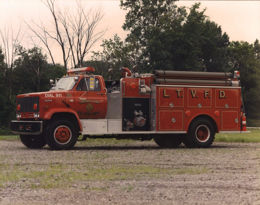 A red fire truck with "L.T.V.F.D." and "Dial 911" on the side, representing the Liberty Township Volunteer Fire Department, is parked on gravel with trees and greenery in the background.