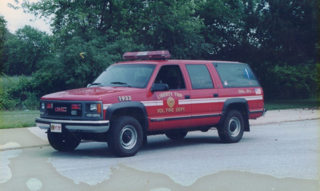 A red Liberty Township Volunteer Fire Department SUV with emergency lights is parked on a paved surface, surrounded by lush greenery and tall trees in the background.