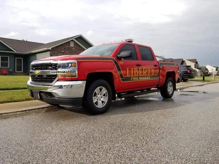 A red pickup truck labeled "Liberty Fire Rescue" from the Liberty Township Volunteer Fire Department is parked on a residential street beside a curb, with houses and cloudy skies in the background.