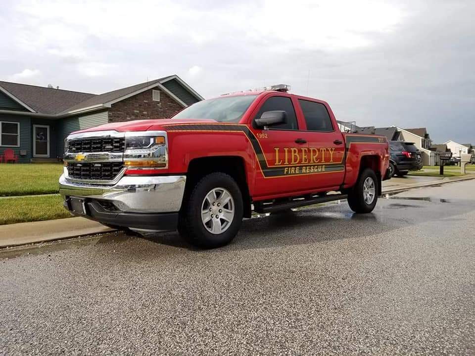 A red pickup truck labeled "Liberty Fire Rescue" from the Liberty Township Volunteer Fire Department is parked on a residential street beside a curb, with houses and cloudy skies in the background.