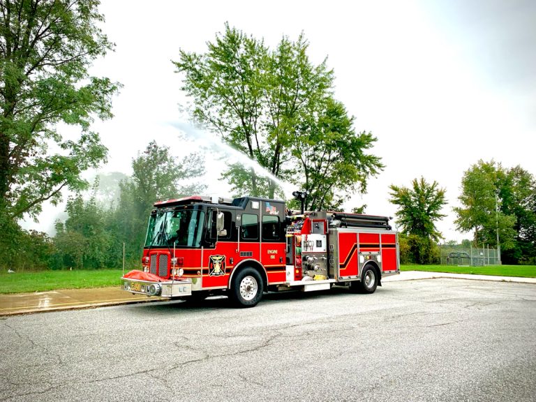 A red Liberty Township Volunteer Fire Department truck is parked on a road, spraying water from its hose. Trees and grass are in the background under a cloudy sky.