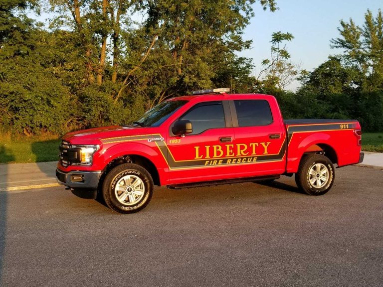 A bright red fire rescue pickup truck with "LIBERTY FIRE RESCUE" and "911" on the side, representing the Liberty Township Volunteer Fire Department, is parked on a paved road with green trees and grass in the background.