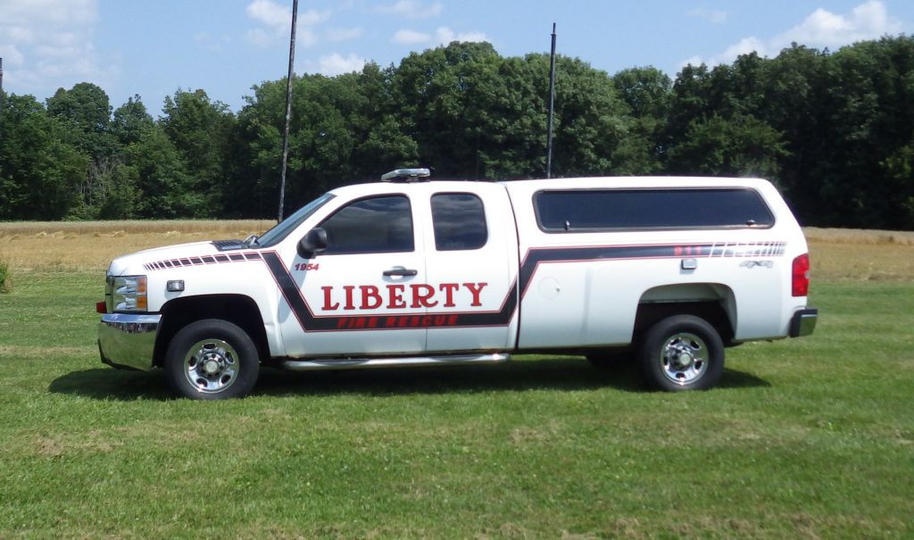 A white pickup truck with "Liberty Township Volunteer Fire Department" and red stripes on the side is parked on grass with trees and a blue sky in the background.