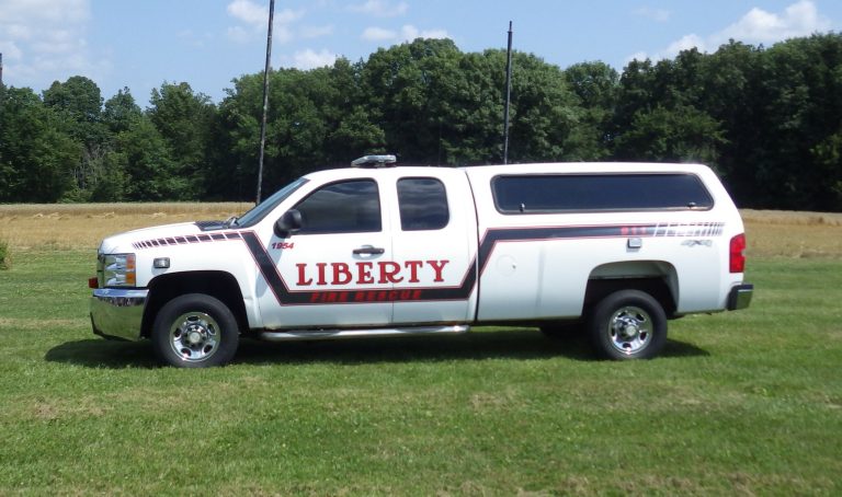 A white pickup truck with "Liberty Township Volunteer Fire Department" and red stripes on the side is parked on grass with trees and a blue sky in the background.