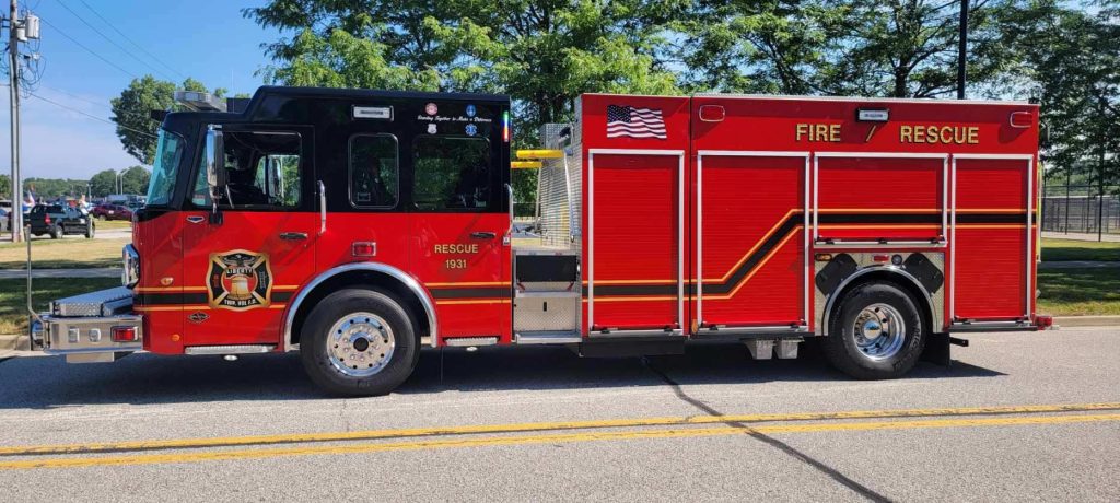 A red Liberty Township Volunteer Fire Department rescue truck with black and gold accents is parked on a street, with trees and a fence in the background. The truck displays an American flag and "FIRE RESCUE" on its side.