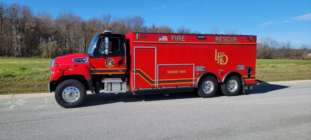 A large red fire rescue truck labeled "Tanker 948" from the Liberty Township Volunteer Fire Department is parked on the street with trees and grass in the background under a clear blue sky.