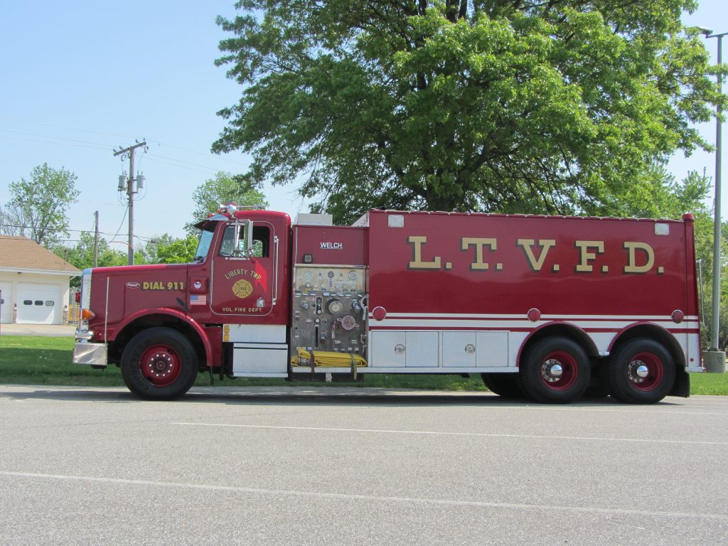 A large red fire truck from the Liberty Township Volunteer Fire Department is parked on a street near grass and trees, with fire equipment visible on its side and "Dial 911" written on the cab door.