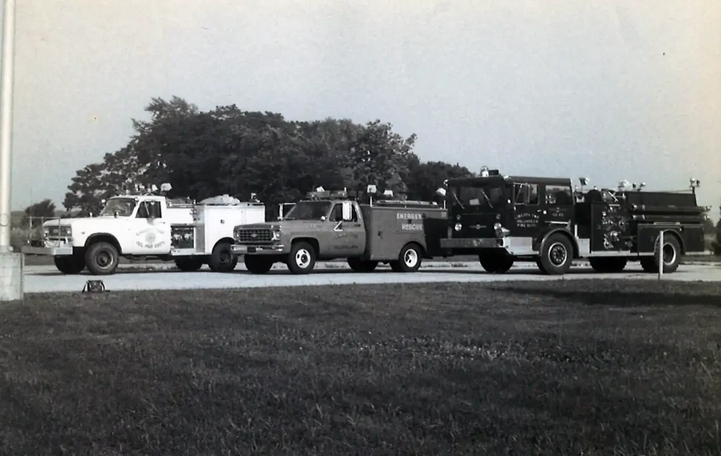 Three vintage emergency vehicles, including two fire trucks and a rescue truck from the Liberty Township Volunteer Fire Department, are parked in a row on a paved surface with grass in the foreground and trees in the background. The image is in black and white.