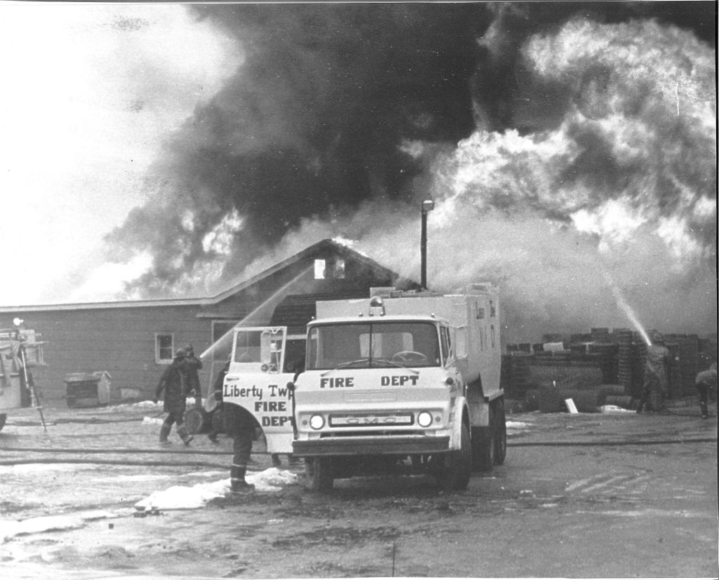 Firefighters from the Liberty Township Volunteer Fire Department spray water on a large building fire as thick smoke billows into the sky. A fire truck is parked in front, with several firefighters visible near the scene.