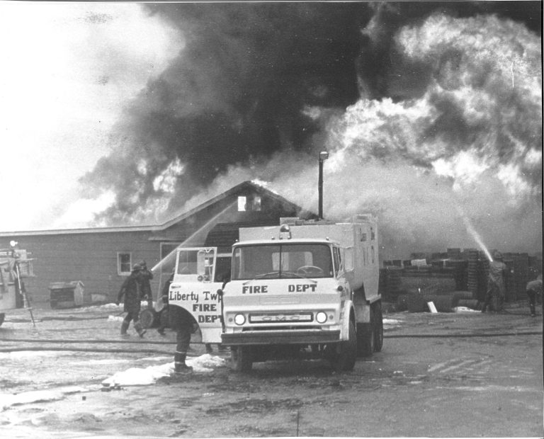 Firefighters from the Liberty Township Volunteer Fire Department spray water on a large building fire as thick smoke billows into the sky. A fire truck is parked in front, with several firefighters visible near the scene.