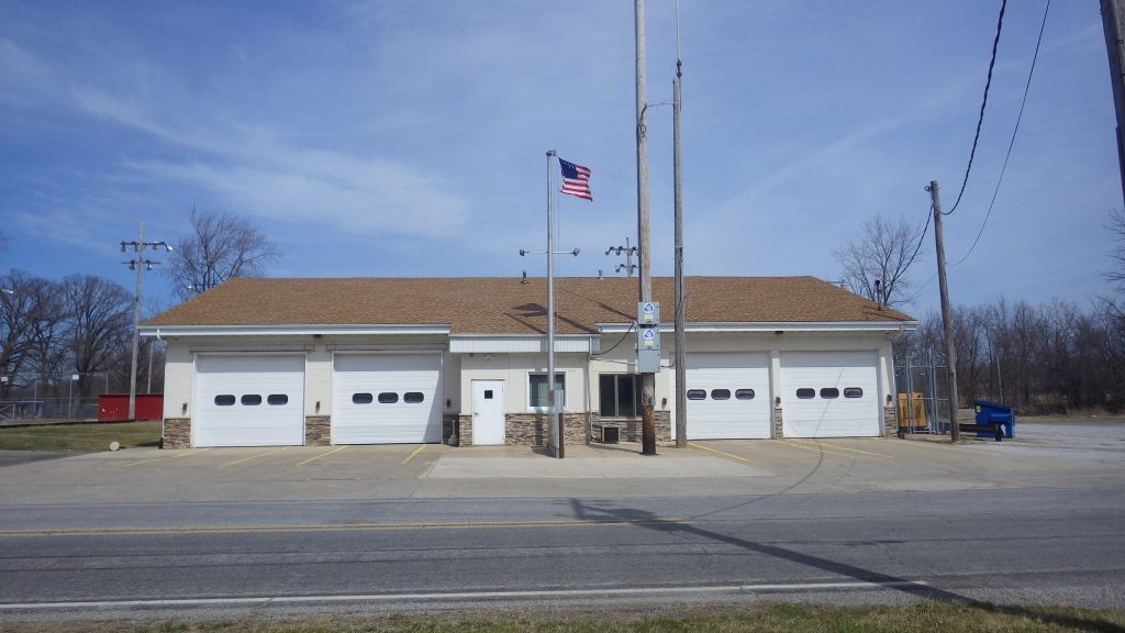 A one-story Liberty Township Volunteer Fire Department station with four garage doors, an American flag on a pole, and a mostly empty parking lot on a clear day. Leafless trees and utility poles are visible in the background.