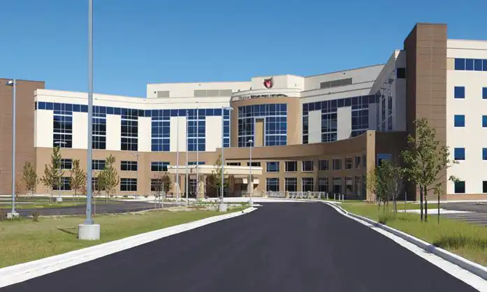 A modern, multi-story hospital building with large windows and a curved entrance sits beside the Liberty Township Volunteer Fire Department, surrounded by a parking lot, trees, and a clear blue sky.