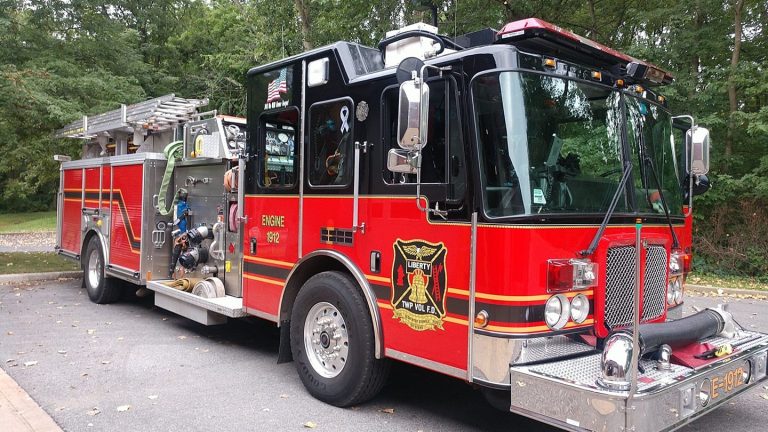 A red and black fire truck with silver accents, marked “Liberty Township Volunteer Fire Department,” is parked on a paved road next to a wooded area, its side displaying firefighting equipment and hoses.