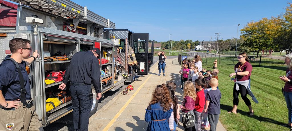 A group of children and adults stand on a sidewalk, watching Liberty Township Volunteer Fire Department firefighters demonstrate equipment from an open fire truck on a sunny day. The scene appears to be an educational visit or demonstration.