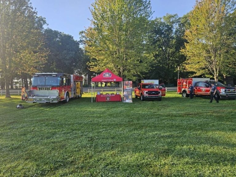 A grassy park with three Liberty Township Volunteer Fire Department vehicles and a red canopy tent set up between them. Firefighters stand near the vehicles, with leafy trees in the background under a clear blue sky.