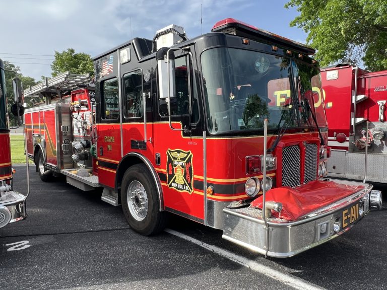 A bright red fire truck with black and gold accents is parked outdoors on a sunny day. The truck is labeled "Engine 1911" and features firefighting equipment and an emblem on the side. Trees and another fire truck are nearby.